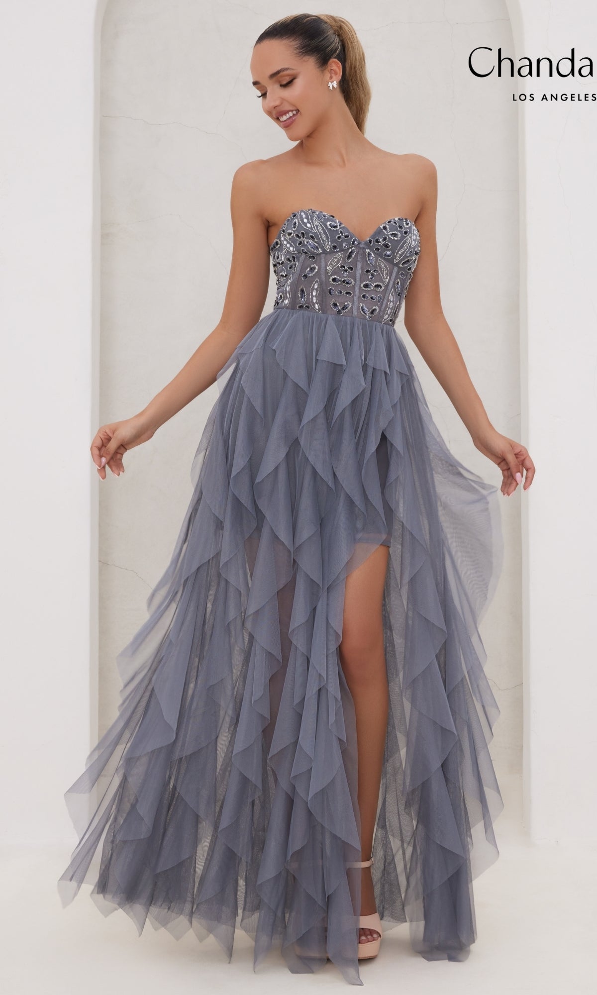 A woman wears the Long Prom Dress 30151 by Chandalier, a strapless slate blue gown with a beaded bodice, layered ruffled tulle skirt, and a thigh-high slit for added allure.